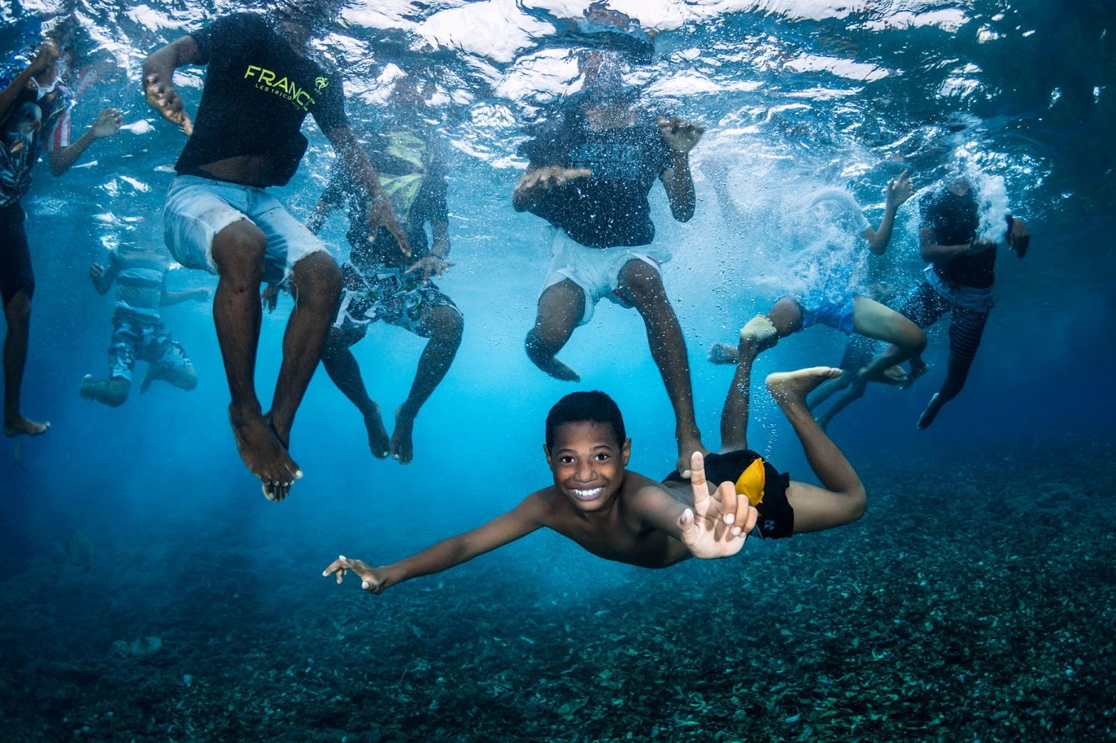Boys swimming. From beneath the water, we see eight boys swimming. Seven of them have their heads above water and we cannot see their faces, but one boy has swum below the water and is floating horizontally, grinning and pointing upwards.